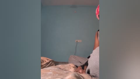 Video of a man lying in bed with a laptop on a teal wall, wearing a gray shirt and beige sheets.
