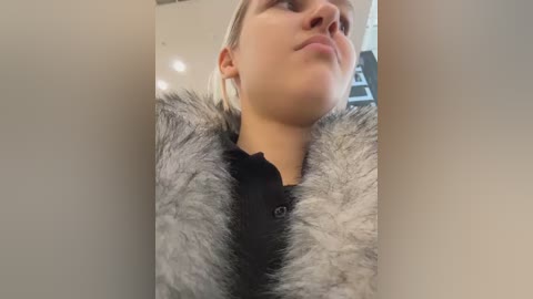 Video of a young Caucasian woman with fair skin and blonde hair, wearing a black shirt underneath a fluffy, gray fur coat. She has a contemplative expression. Background is a blurred, indoor setting.