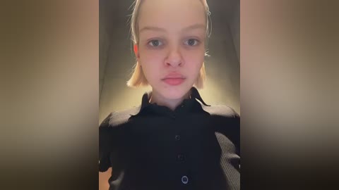 Video of a young girl with pale skin and straight, light blonde hair. She wears a black, button-up blouse with a subtle pattern. The background is blurred, creating a soft, intimate atmosphere.