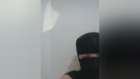 Video of a person in a black mask and hoodie, partially visible, standing in a dimly lit, plain white room with a large, blurry white object in the background.