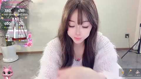 A video of an East Asian woman with long black hair, wearing a white knitted sweater, looking down in a minimalist room with a plant and a digital speedometer overlay.
