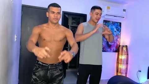 Video of two shirtless, muscular men in a modern room with colorful wall art, one in black pants, the other in a grey tank top.