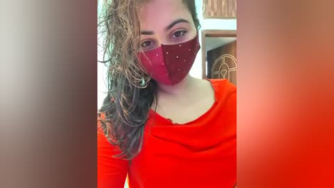 Media: Video of a woman with wet, wavy brown hair, wearing a red face mask and a red long-sleeve top, indoors, blurred background.