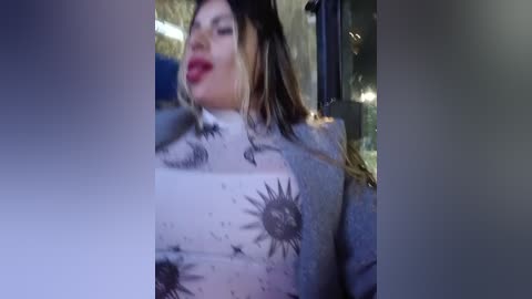 A video captures a woman with fair skin and long, wavy hair, wearing a grey blazer with sunflower patterns. She has red lipstick and is smiling. The background is blurry, suggesting an indoor setting.