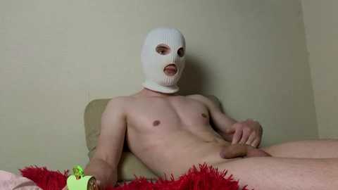 Video of a nude, fair-skinned man with a white mask, seated on a bed with a red blanket. He has a flaccid penis and small breasts. Background is a plain beige wall.