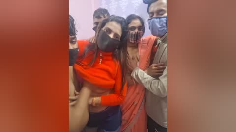 A video shows a group of five people, four wearing face masks, huddled closely. The woman in the center, with long dark hair, wears a bright orange top. The background is blurry.