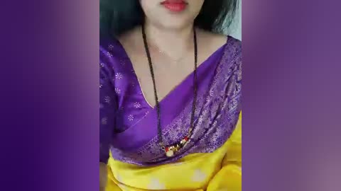 A video of an Indian woman wearing a vibrant purple saree with gold embroidery, yellow blouse, and a black beaded necklace, her lips painted red, against a purple gradient background.