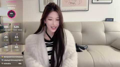 A video of an East Asian woman with long black hair, fair skin, and a slim figure, wearing a white fur coat over a black and white striped shirt, smiling. She sits on a beige sofa in a modern living room with framed art on the wall.