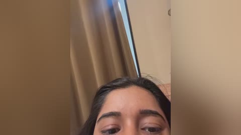 Video of a young woman with medium skin tone and long, dark hair, partially visible from the waist up. She is looking directly at the camera with focused eyes. Background features a beige wall and a vertical window with light streaming in.