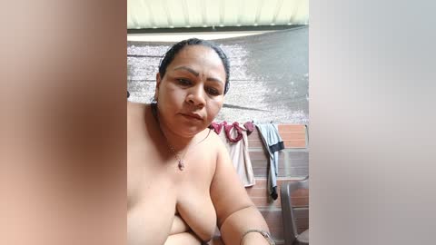 A candid video of a topless, middle-aged Asian woman with medium skin tone, medium-sized breasts, and a neutral expression, standing in a rustic bathroom with a tiled wall and wooden bench.