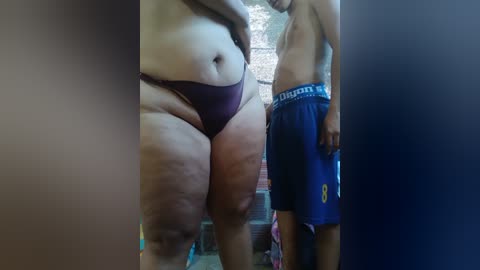Video: Two overweight individuals in a bathroom. A woman with fair skin and a large body, wearing maroon panties, stands next to a shirtless man with a light brown complexion, wearing blue shorts.