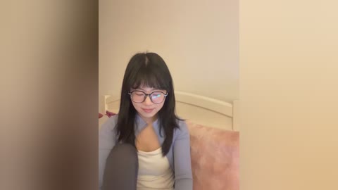 Video of an Asian woman with long black hair, wearing glasses, a light blue sweater, and white top, sitting on a bed with a beige blanket, smiling softly.