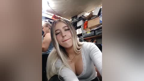 Video of a young, light-skinned woman with long, blonde hair and a pierced nose, wearing a low-cut white top, lying in a messy storage room.