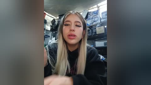 Video of a young woman with long, straight blonde hair and light skin, wearing a black top, sitting in a cluttered, dimly lit room with shelves filled with electronics and cables.