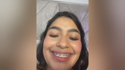 Video of a young woman with medium brown skin, long black hair, and braces, smiling broadly. She has light pink eyeshadow and blush, and is wearing a white top. Background features a light beige, patterned couch.