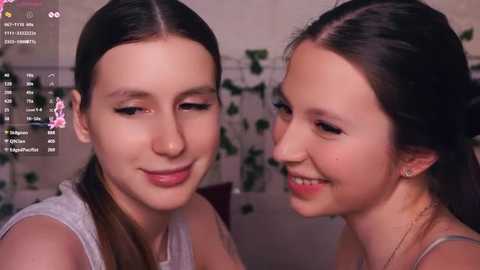 Video of two young women with light skin, straight dark hair, and wearing casual outfits, smiling, in a cozy indoor setting with green plants and a dimly lit background.