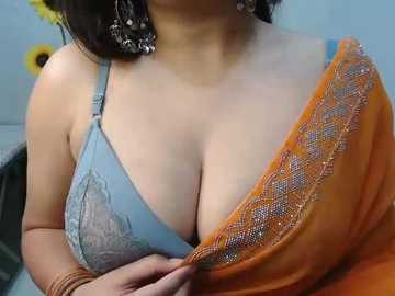 Video of a woman with medium skin tone, wearing a light blue lace bra with medium-sized breasts. She has an orange sari partially draped over her shoulder, adorned with intricate embroidery.