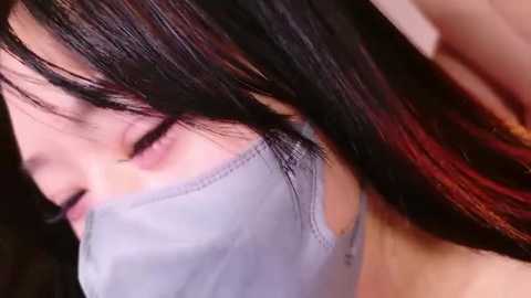 A close-up video of a person with long, straight black hair and a light blue face mask, partially obscured by the camera angle. The background is blurred, focusing attention on the mask and hair.