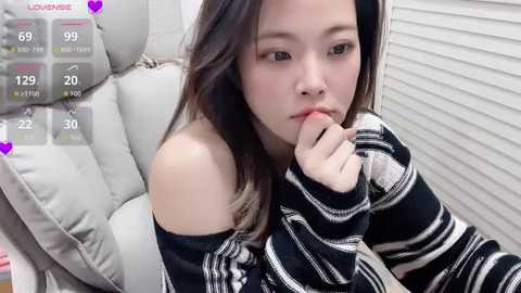 Video of a young Asian woman with long, dark hair, wearing a striped sweater, sitting in a car seat, looking pensive, with a heart overlay.