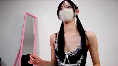 Video of an Asian woman in a black maid outfit with white lace trim, face mask, and white headband, standing in a minimalist room with a pink mirror and white walls.