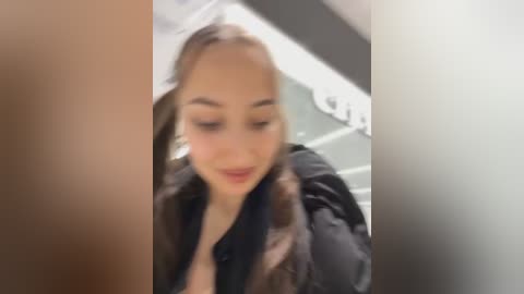 A blurred video of a young woman with light skin, brown hair, and wearing a black jacket, captured indoors in a dimly lit setting.