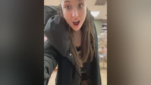 Video of a young woman with long brown hair, wearing a black coat, looking surprised and raising her arm in a supermarket aisle.