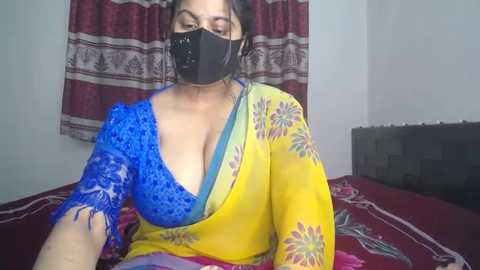 Video of a South Asian woman with medium skin tone, wearing a blue lace blouse and yellow sari with floral patterns, black face mask, and black hair tied back. She is on a red bed with a floral-patterned blanket.