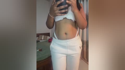 A video of a young woman with medium brown skin, wearing a white lace bra and white pants, taking a mirror selfie in a bedroom.