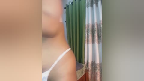 Video of a blurred person wearing a white tank top, standing near a window with green curtains and a wooden table.