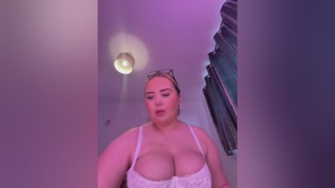 Media: A video of a plus-size, fair-skinned woman with large breasts wearing a white lace bra, standing in a dimly lit room with purple lighting. She has glasses on her head and is looking downwards.