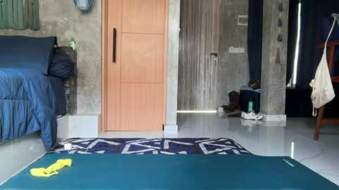 Video of a small, cluttered bedroom with a blue bed, wooden door, and concrete walls. A green yoga mat, a yellow item, and a white towel are on the floor.