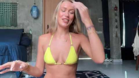 Media: Video of a smiling, blonde woman in a yellow bikini, adjusting her hair, in a dimly lit, modern room with concrete walls and blue furnishings.