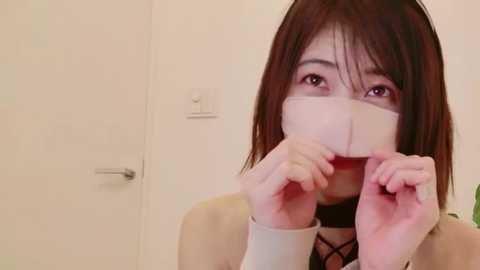 Media: A video of an Asian woman with shoulder-length brown hair, wearing a beige coat, holding a piece of paper over her mouth, in a plain, beige-walled room with a door and light switch visible.