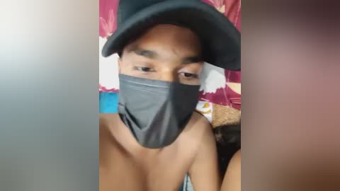 Media: Video of a topless person with light brown skin, wearing a black face mask, a black hat, and a colorful floral-patterned pillow.