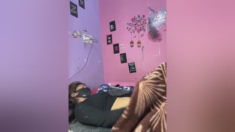 Video of a woman in black, lying on a bed, wearing a mask, in a room with purple and pink walls adorned with art and medical equipment.