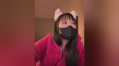 Media: Video of a young Asian woman with long black hair, wearing a pink hoodie, black face mask, and cat ears, standing in a dimly lit room with beige walls.