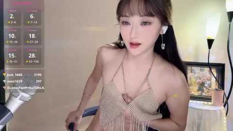 A young East Asian woman with long black hair, wearing a beaded halter top, sits in a modern room, adorned with a computer screen displaying streaming statistics.