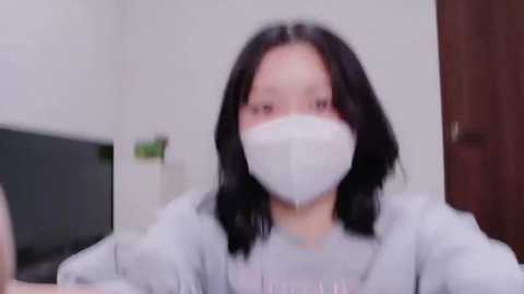 A blurred video of an Asian woman with shoulder-length black hair, wearing a white mask and light grey sweater, standing in a dimly lit room with white walls and a wooden door.