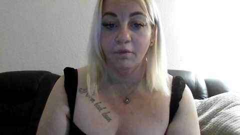 Video of a middle-aged, fair-skinned woman with shoulder-length blonde hair, wearing a black bra and necklace. She has tattoos on her chest and neck. The background is a plain, light-colored wall and a dark sofa.