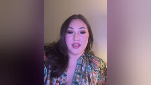 Media: Video of a young Asian woman with long black hair, wearing a colorful floral robe, smiling against a plain beige wall.