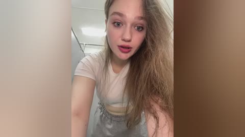 Media: Video of a young, light-skinned woman with long, wavy blonde hair, wearing a white t-shirt and gray sweatpants, leaning forward, mouth slightly open, in a dimly lit bathroom with beige walls and a shower curtain.