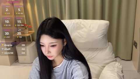 Video of a young Asian woman with long black hair, wearing a light gray t-shirt, sitting on a white cushion, indoors with beige curtains and a table in the background.