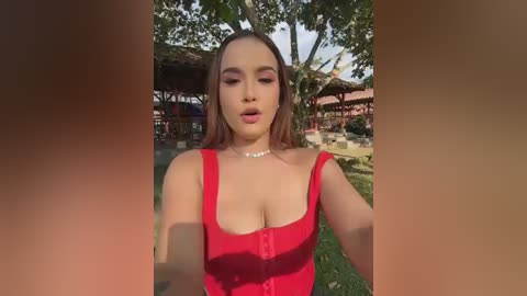 Video of a young Asian woman with light skin and long brown hair, wearing a red off-shoulder top, smiling outdoors near a wooden hut with green trees.