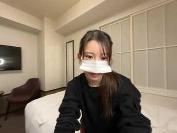 A video of an East Asian woman with long dark hair, wearing a white surgical mask, black shirt, and seated on a bed in a sparsely furnished, beige-toned room with a window and TV.