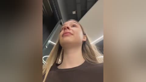 Video of a young Caucasian woman with straight, blonde hair, wearing a black top, standing in a modern, well-lit indoor space with a glass railing and dark ceiling.