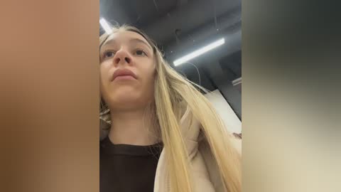 A video of a young Caucasian woman with long, straight blonde hair and light skin, wearing a black top and beige jacket, taken from a low angle, looking up. Background shows industrial ceiling with fluorescent lights and pipes.