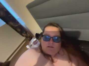 A candid video of a fair-skinned woman with glasses and auburn hair, lying on a bed, wearing only blue sunglasses, with a small white cat near her head.
