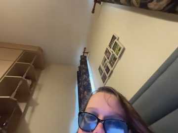 A video of a person wearing black glasses and a light-colored sweater, standing in a narrow, beige-walled room with a model train track on the floor and a shelf with a dog on it in the background.