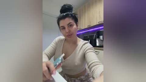 A video of a young woman with dark hair in a bun, wearing a beige crop top and patterned skirt, applying makeup in a modern kitchen with wooden cabinets and purple LED strip lighting.