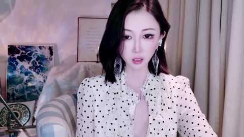 Video of an East Asian woman with straight black hair, wearing a white polka-dot blouse, large silver earrings, and light makeup, sitting indoors with a modern, abstract ocean-themed painting in the background.